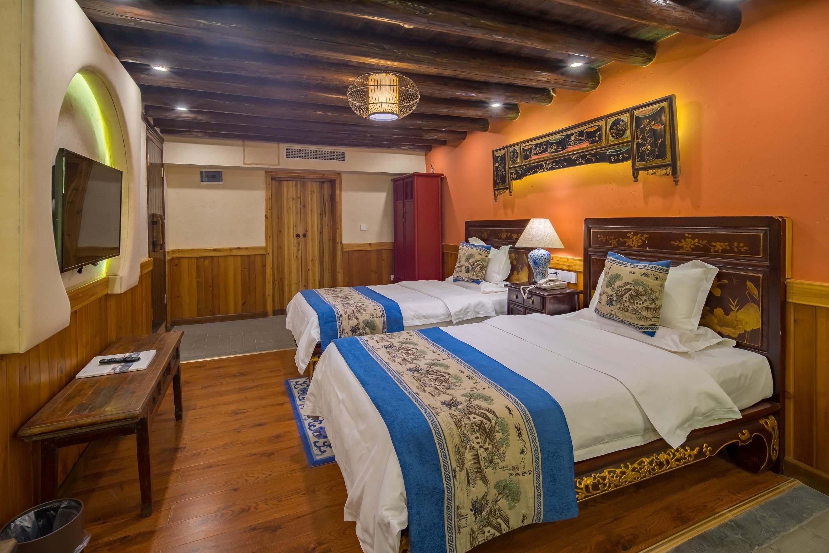 A hotel room with two beds, wooden flooring, a television, a desk, and a red cabinet.