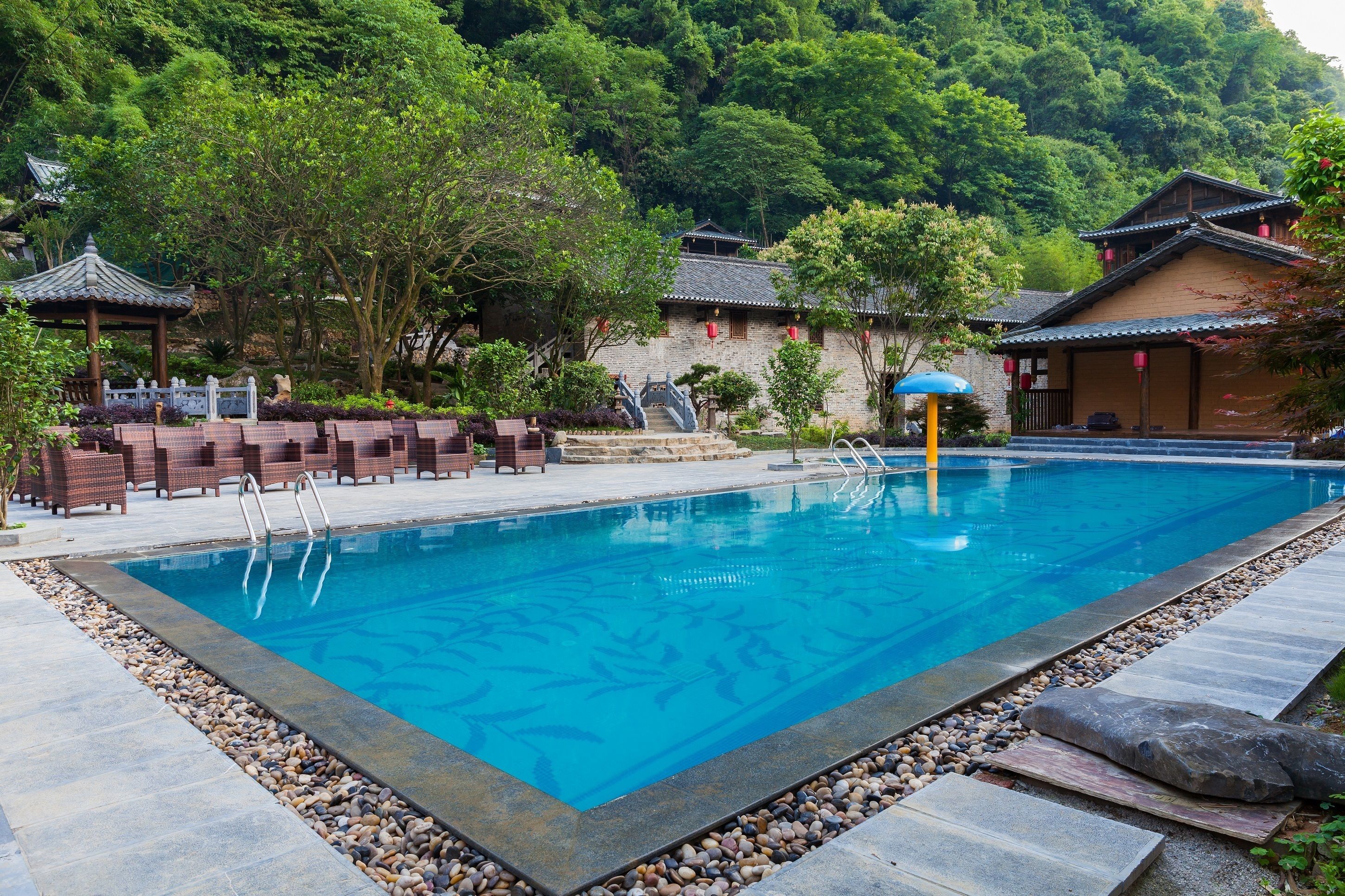 Una piscina con borde de piedra, rodeada de arquitectura tradicional y abundante vegetación.