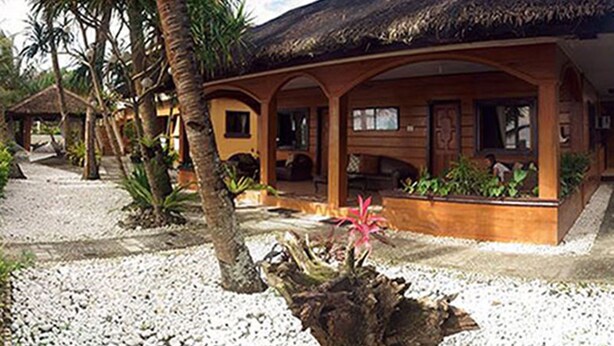 A wooden building with a thatched roof, surrounded by palm trees and a gravel path. - Pandan Beach Resort (Pandan)