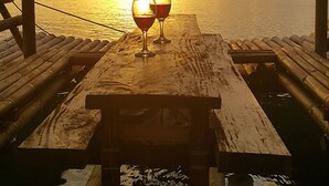 A wooden table with two wine glasses under a wooden pergola with draped fabric, overlooking a sunset over the sea. - Pandan Beach Resort (Pandan)