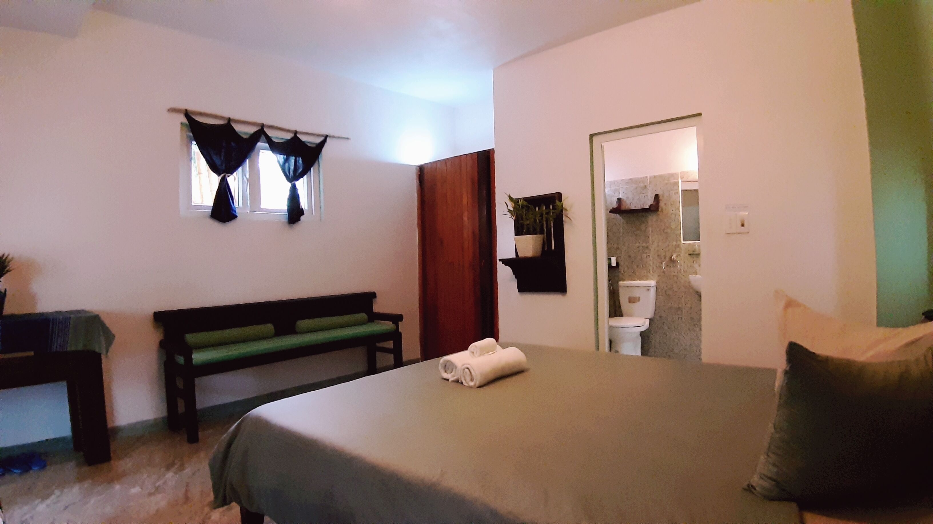 A bedroom with a bed, a bench, a small table, a window with curtains, and a bathroom visible through an open door.