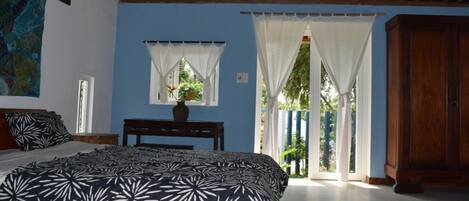 A bedroom with a bed, a nightstand, a window with curtains, a wooden ceiling, and a view of the outside.