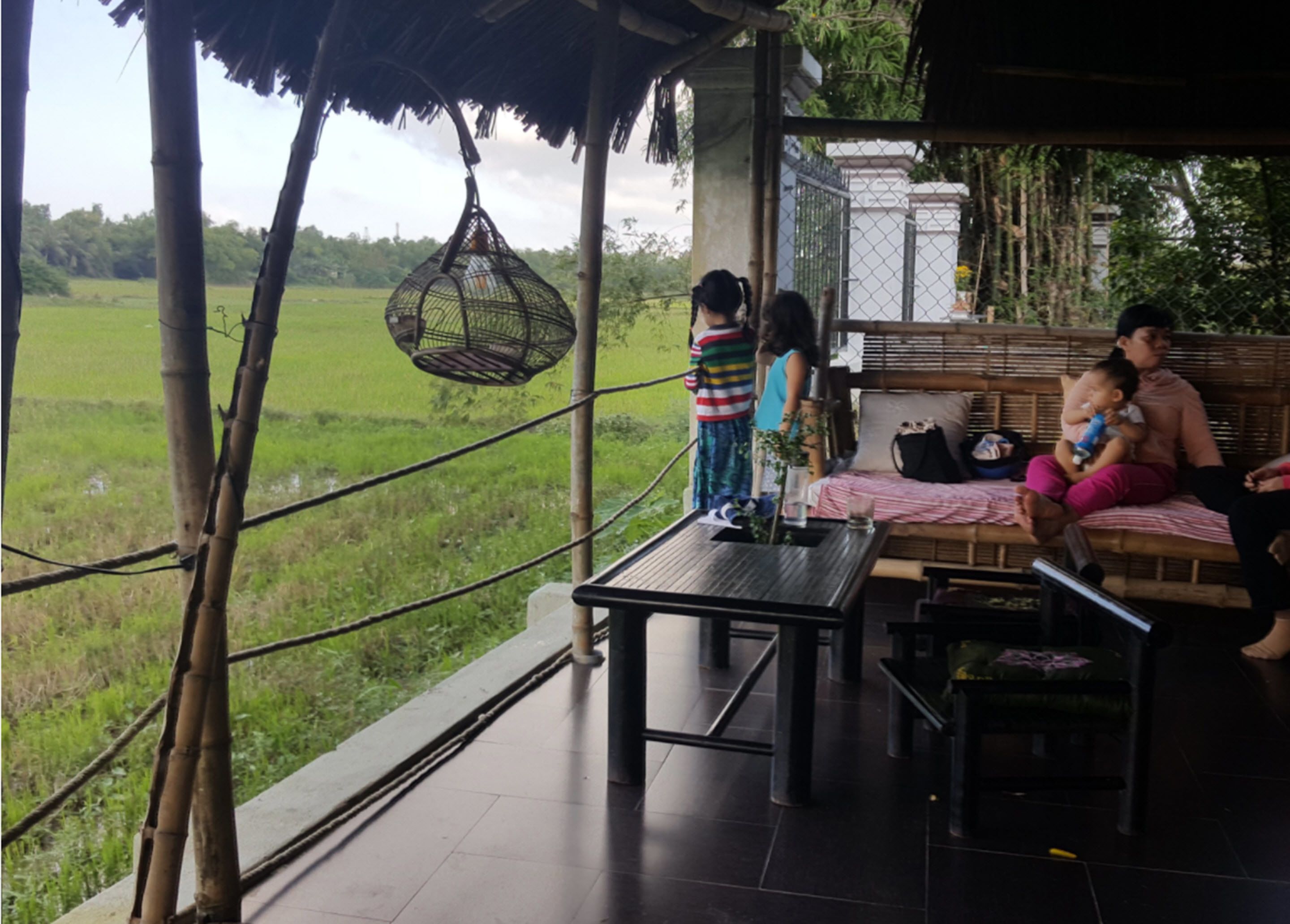 Un espacio de descanso cubierto al aire libre con vista a un campo verde. Hay una mesa con un vaso y una planta, además de una silla colgante.
