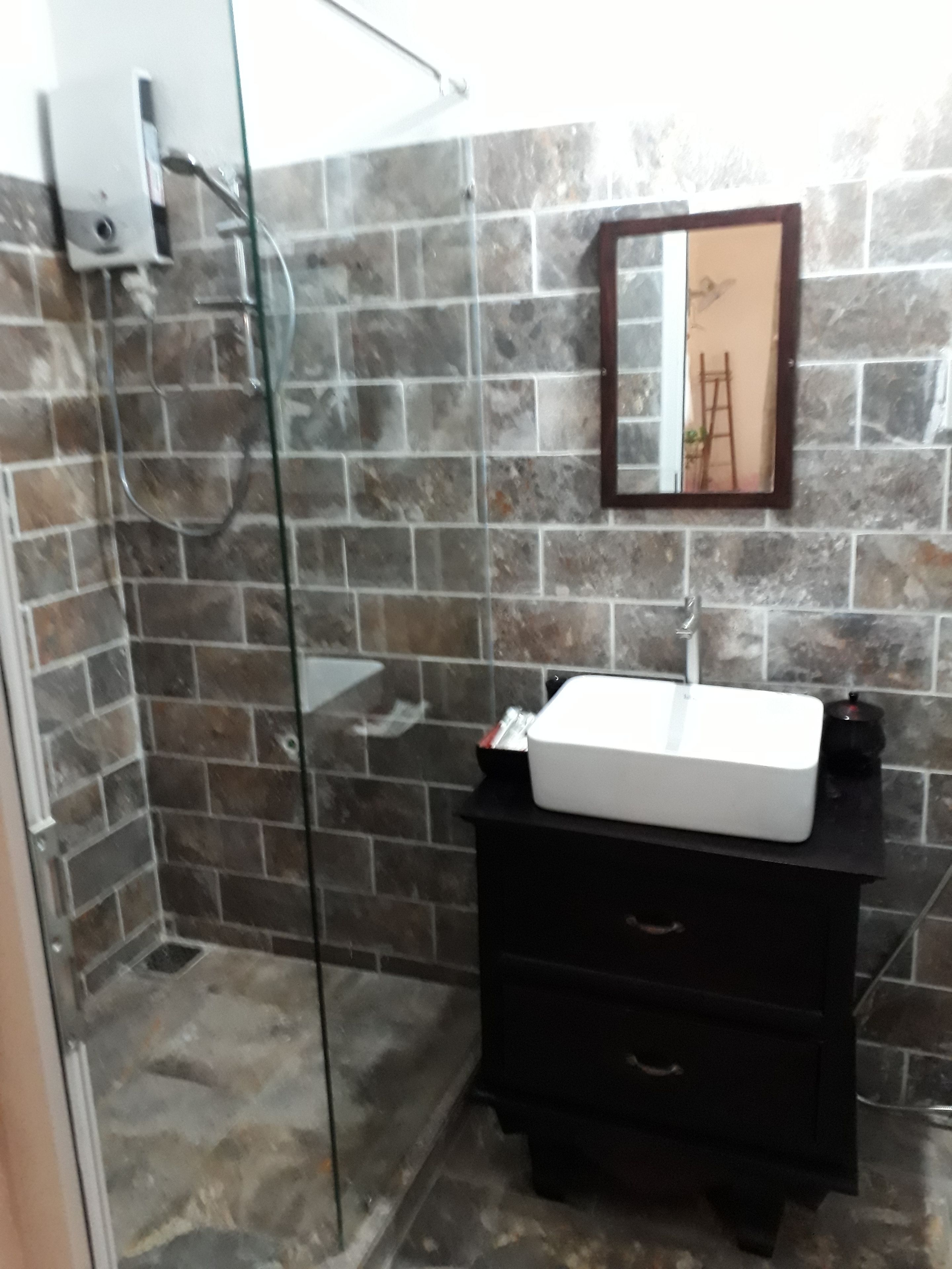 A bathroom with a glass shower enclosure, a white sink on a black vanity, and a stone-tiled wall.