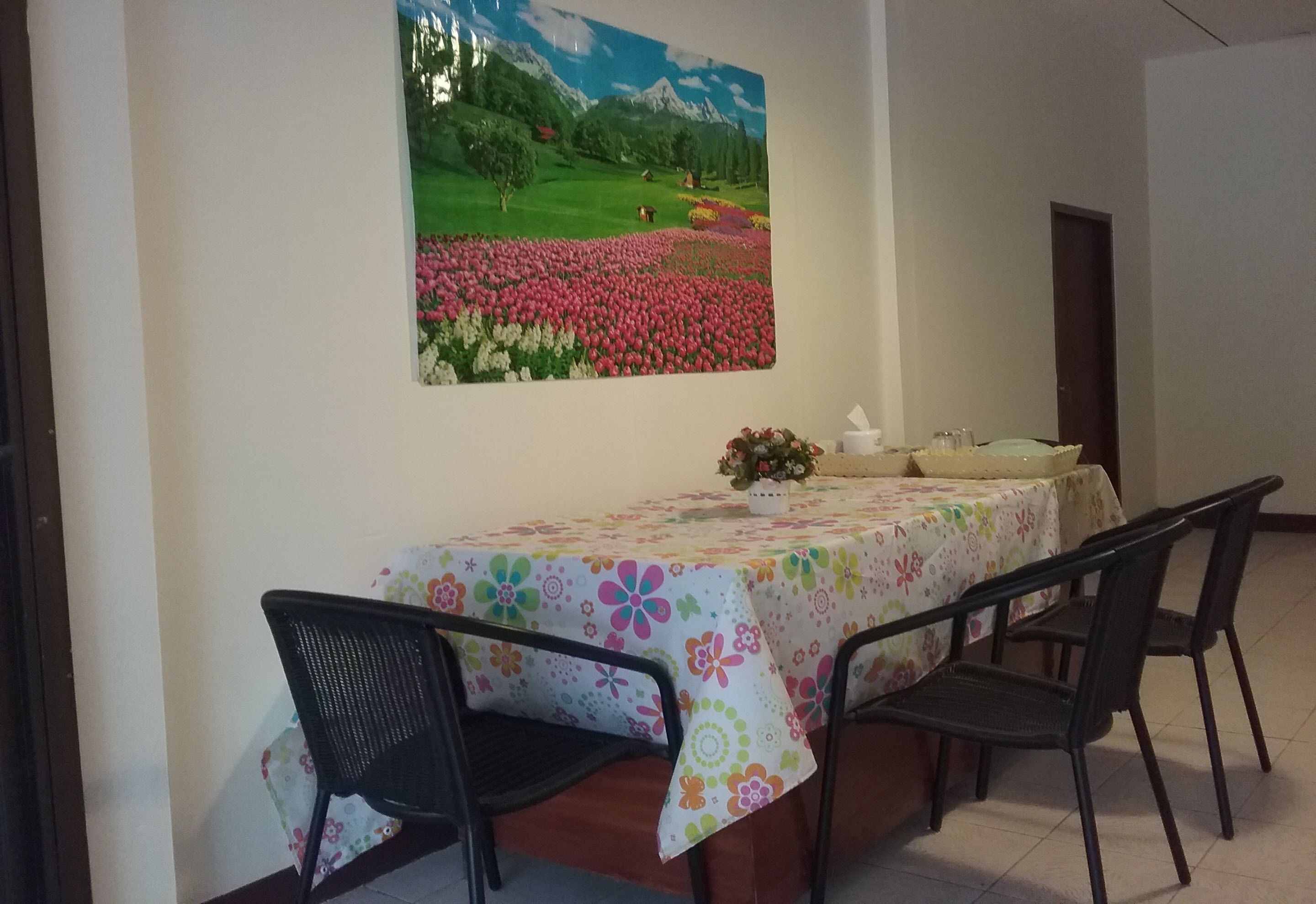 A dining area with a table set for four, chairs, a floral tablecloth, and a painting of a field with flowers and mountains.