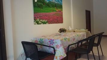 A dining area with a table set for four, chairs, a floral tablecloth, and a painting of a field with flowers and mountains.