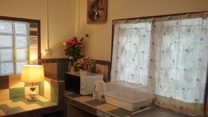 Una kitchenette pequeña con microondas, tostadora y lavamanos. Se ve una ventana con cortina estampada y un florero con flores en la encimera.