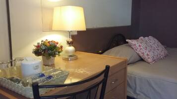 A hotel room with a wooden desk, a lamp, a flower vase, a bed with floral pillows, and a mirror on the wall.