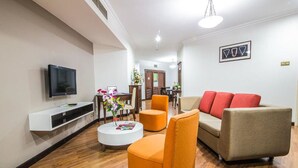 Ein modernes Wohnzimmer mit einem Flachbildfernseher, orangefarbenen und beigen Sofas, einem weißen Couchtisch und einem Essbereich im Hintergrund.
