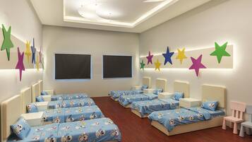 Quarto infantil com várias camas, cada uma com colcha azul estampada com personagens de desenhos animados, cabeceiras brancas e mesas de cabeceira.