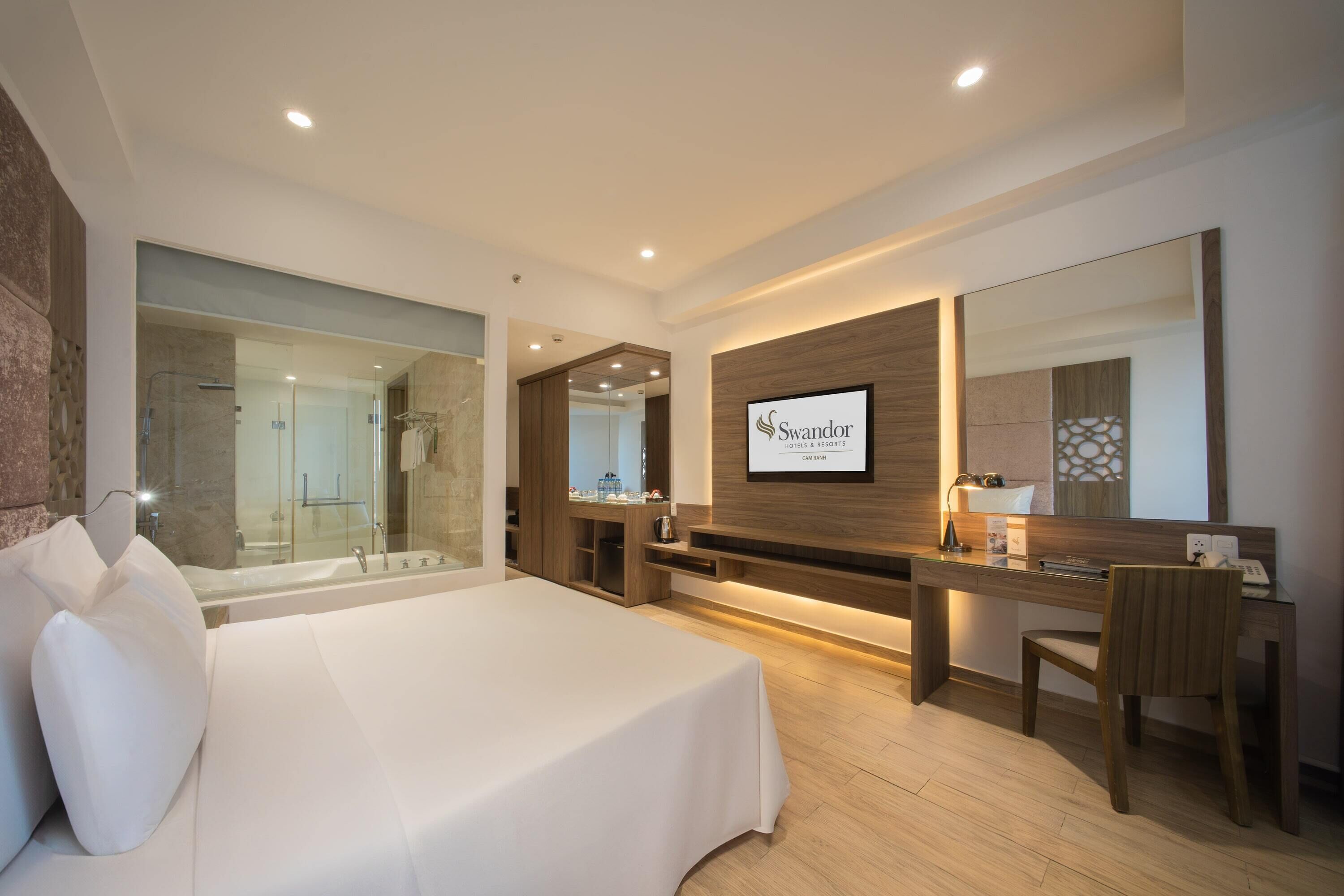 Quarto de hotel moderno com uma cama grande, uma escrivaninha, uma cadeira, área de chuveiro e uma televisão montada exibindo o nome do hotel.
