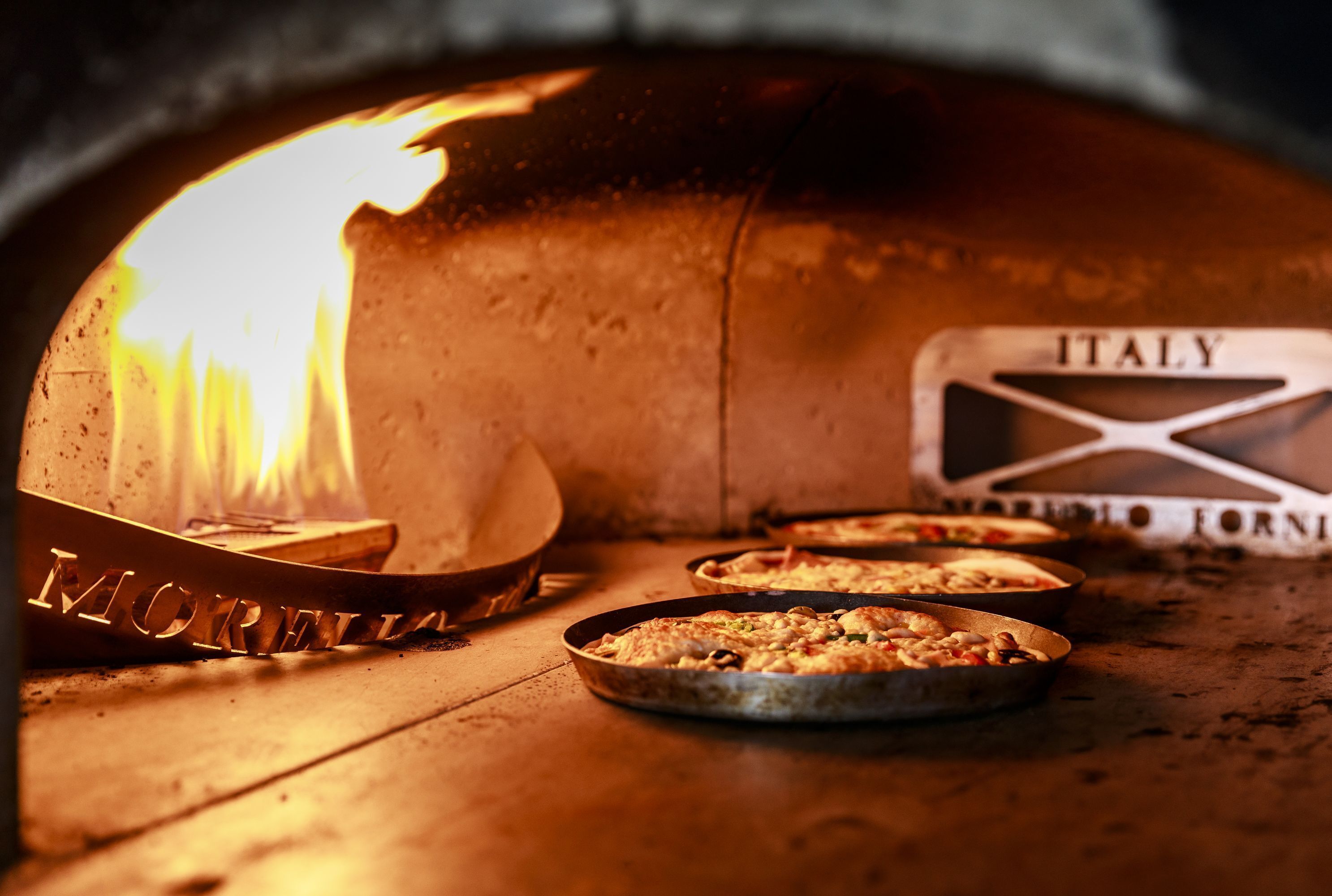 A wood-fired oven with pizzas inside, a sign reading "ITALY," and a metal tray with the word "MORELLA."
