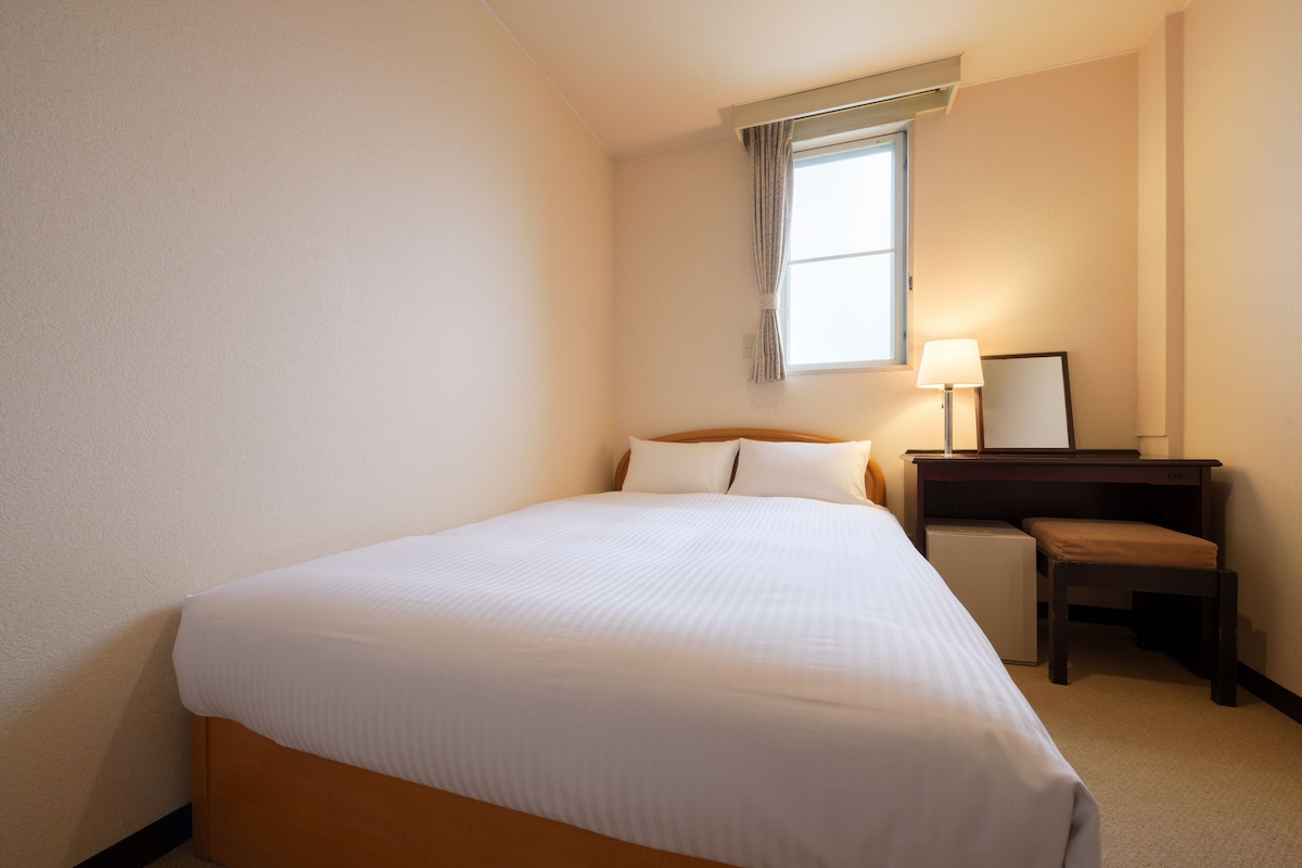Une chambre d’hôtel avec un lit, un bureau, une chaise, une lampe et une fenêtre.