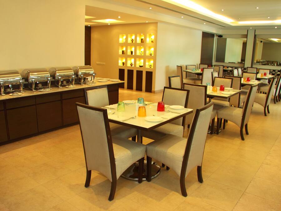 A hotel dining area with multiple tables and chairs, a buffet line with food dispensers, and a wall of illuminated lights.