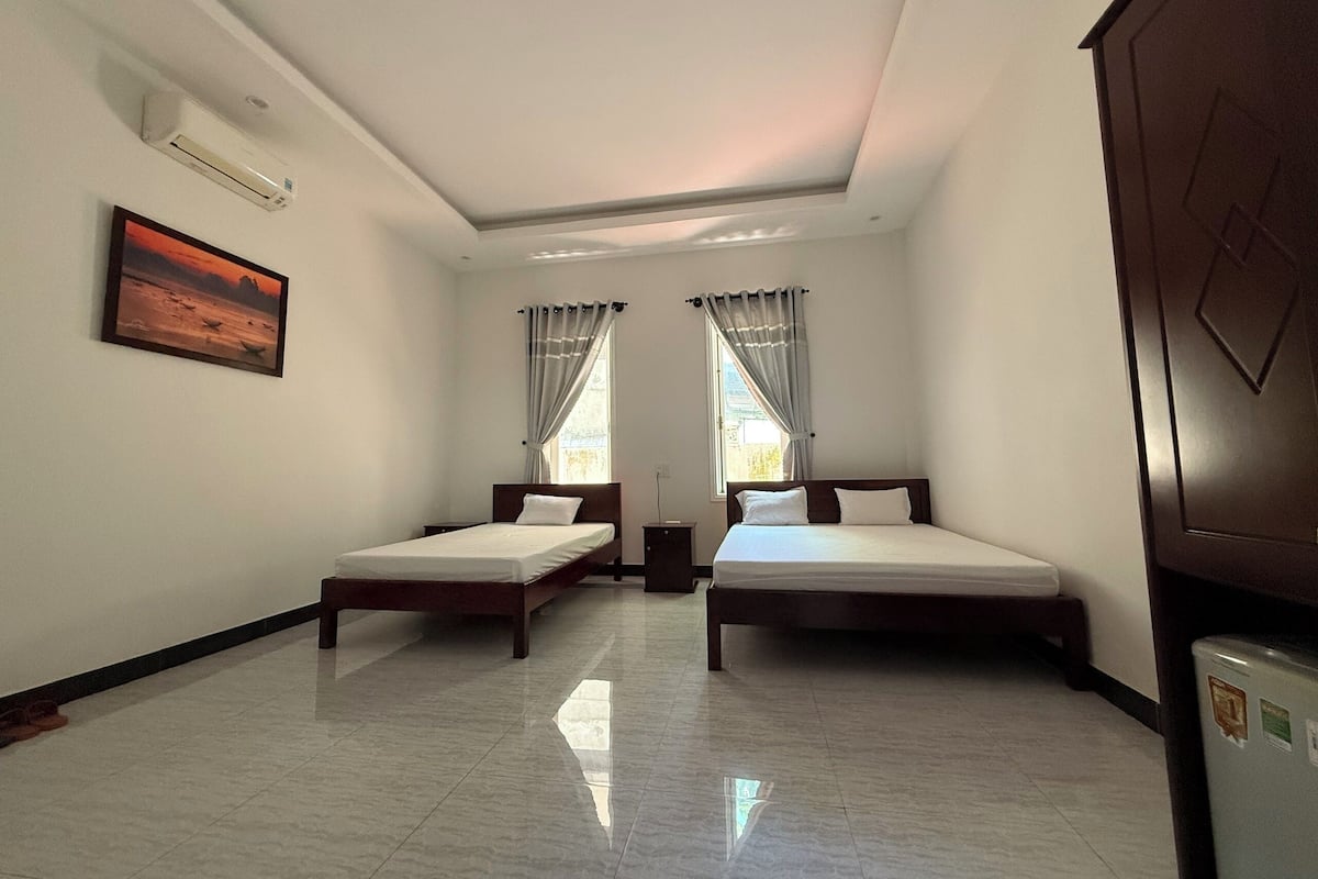 A room with two single beds, a ceiling fan, a wall-mounted air conditioner, a framed picture, and a window with curtains.