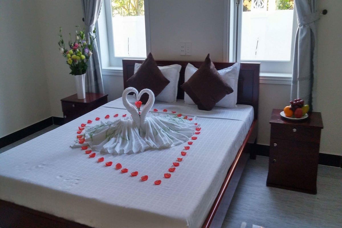 A neatly made bed with a swan-shaped decoration made of white fabric and red petals, a vase of flowers on a bedside table, and a fruit bowl on another table.