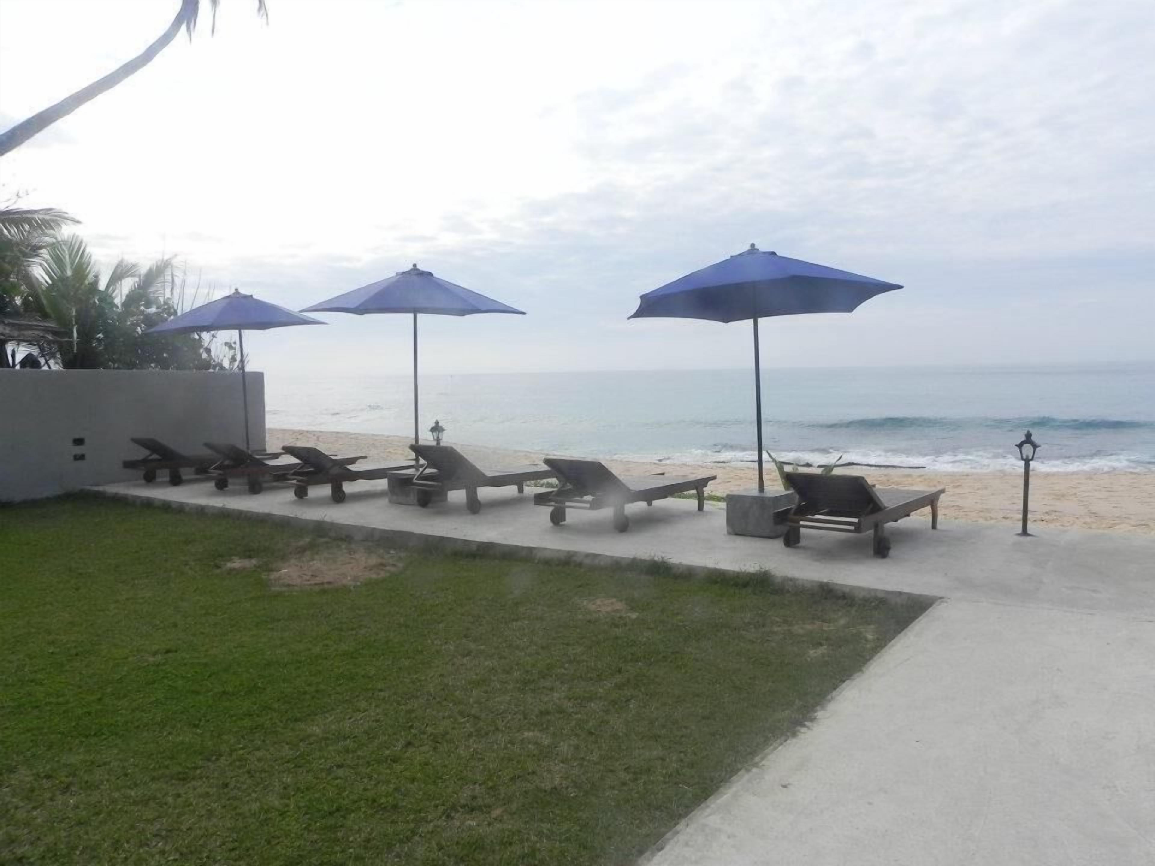 Een terras aan het strand met ligbedden en parasols, een betonnen pad en uitzicht op de oceaan.