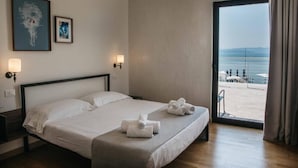 Een hotelkamer met een bed, twee wandlampen, een groot raam met uitzicht op zee, en twee ingelijste kunstwerken aan de muur.