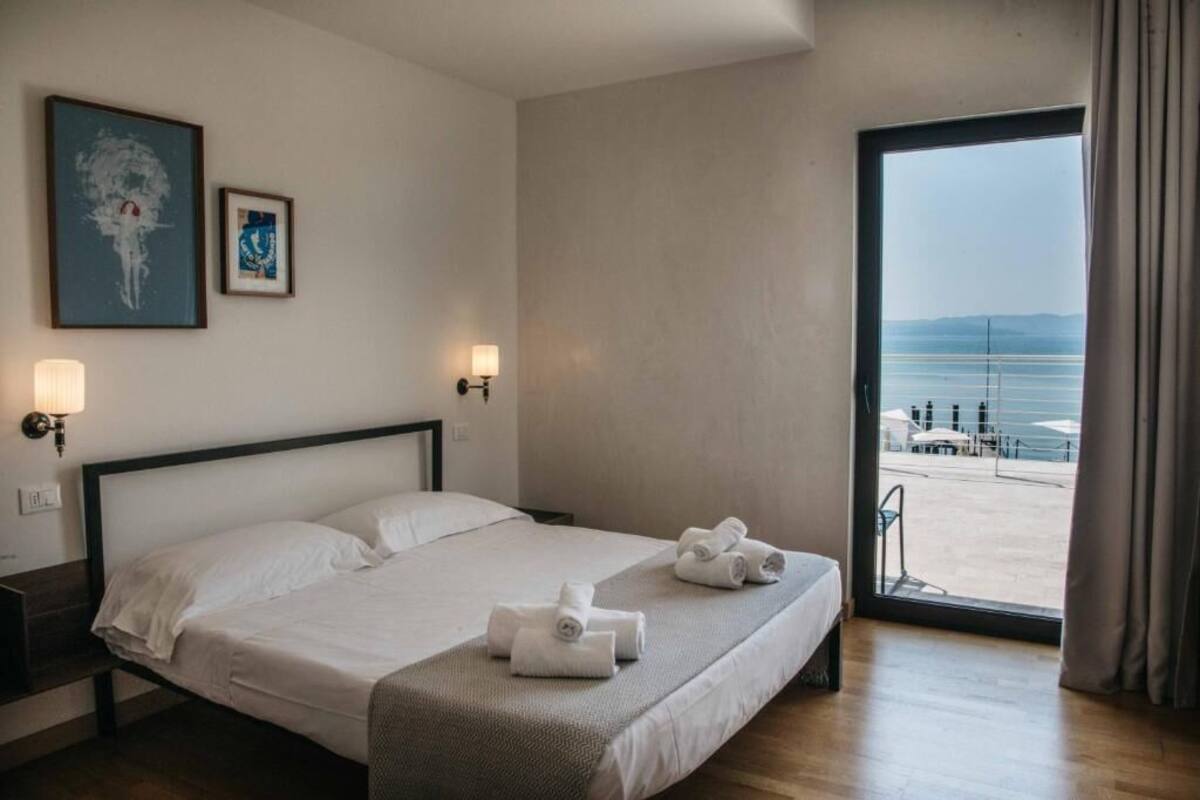 Een hotelkamer met een bed, twee wandlampen, een groot raam met uitzicht op zee, en twee ingelijste kunstwerken aan de muur.