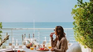 Iemand zit aan een tafel met uitzicht op zee, een plant en een dienblad met fruit en een fles.