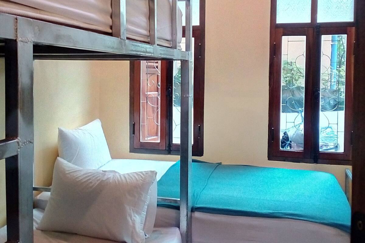 A dormitory room with bunk beds, a window with a metal grill, and an air conditioning unit on the wall.