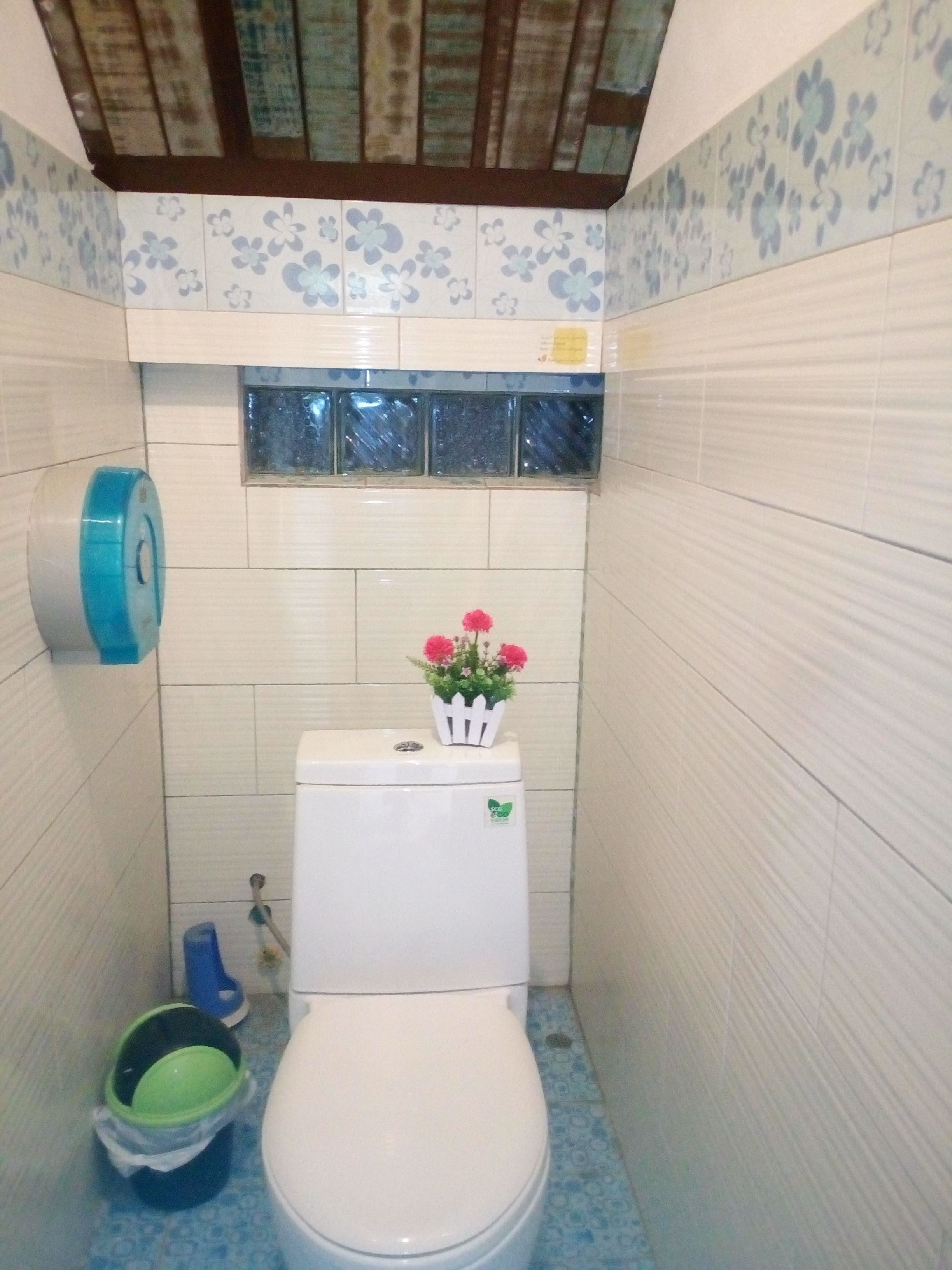 A small bathroom with a toilet, a blue patterned floor, a window, and a hand sanitizer dispenser.