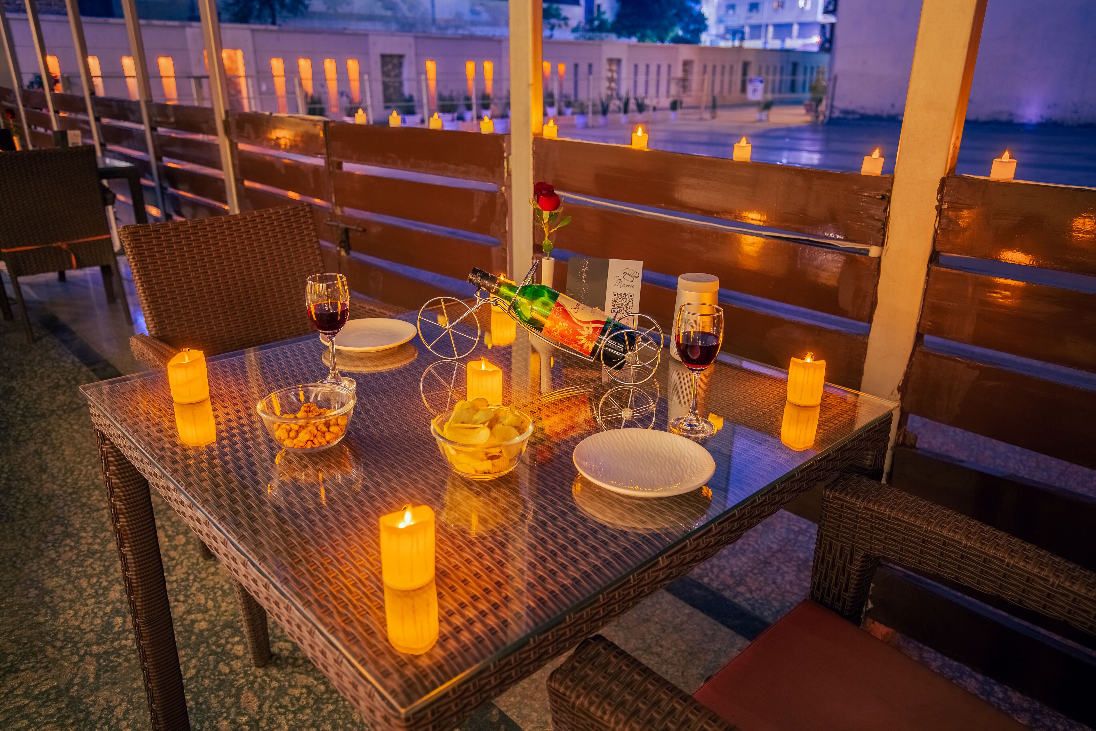 Una mesa dispuesta para dos con velas, una botella de vino, un cuenco de aperitivos y una rosa.