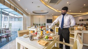 A hotel dining area with tables set for breakfast, a waiter serving, and a large window offering a view. - Rosewood Apartment Hotel - Gurgaon (Gurugram)