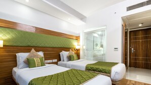 A hotel room with two beds, wooden headboards, green and white bedding, a glass shower, and a wooden door. - Rosewood Apartment Hotel - Gurgaon (Gurugram)