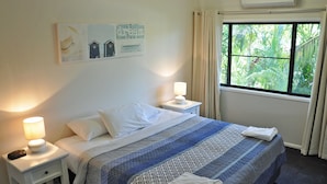 A neatly made bed with a patterned bedspread, two bedside lamps, a nightstand, a window with curtains, and a wall with a framed picture. - Angelwaters (North Ward)