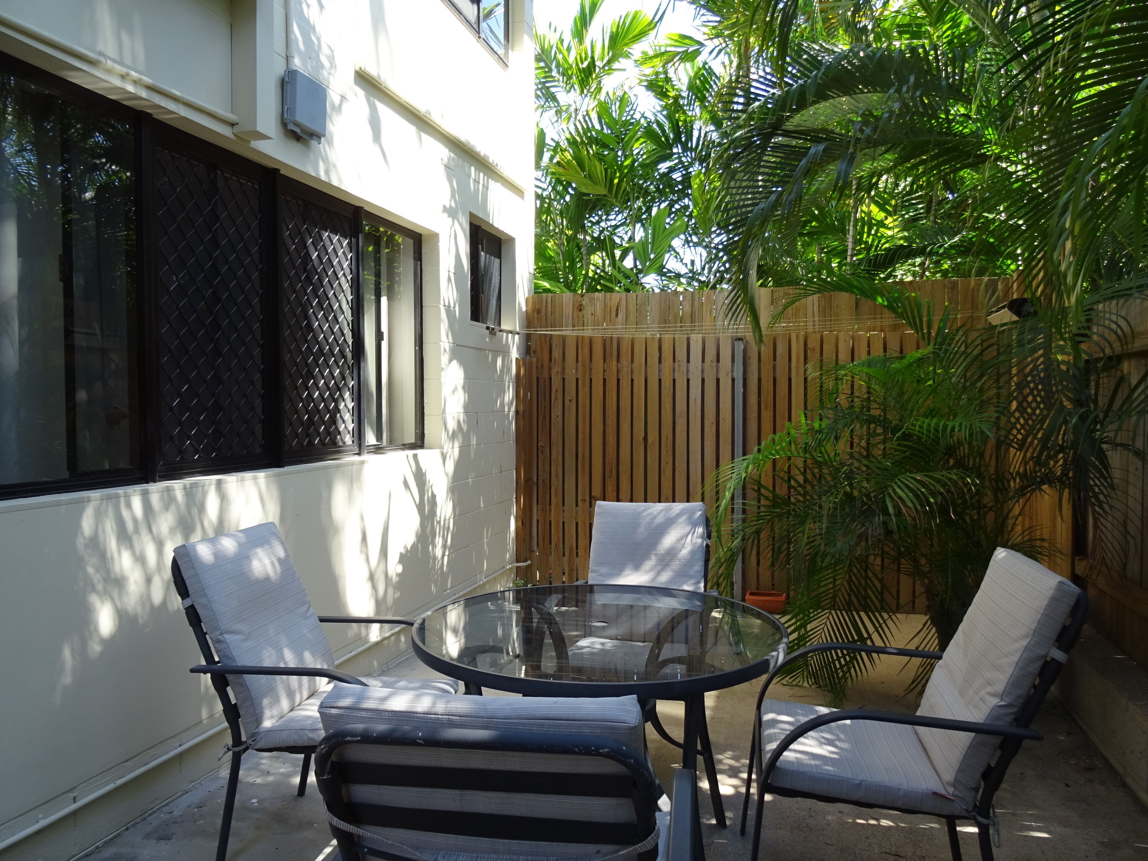 A glass-top table with four chairs, a wooden fence, and a house with white walls and a window with a metal screen.