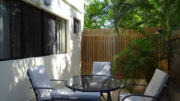 A glass-top table with four chairs, a wooden fence, and a house with white walls and a window with a metal screen.