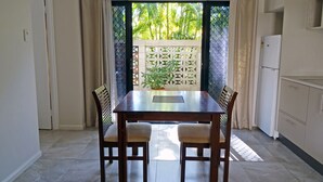 A dining area with a table and four chairs, an air conditioning unit above, a view of greenery through a window, and a kitchen area with cabinets and a refrigerator. - Angelwaters (North Ward)