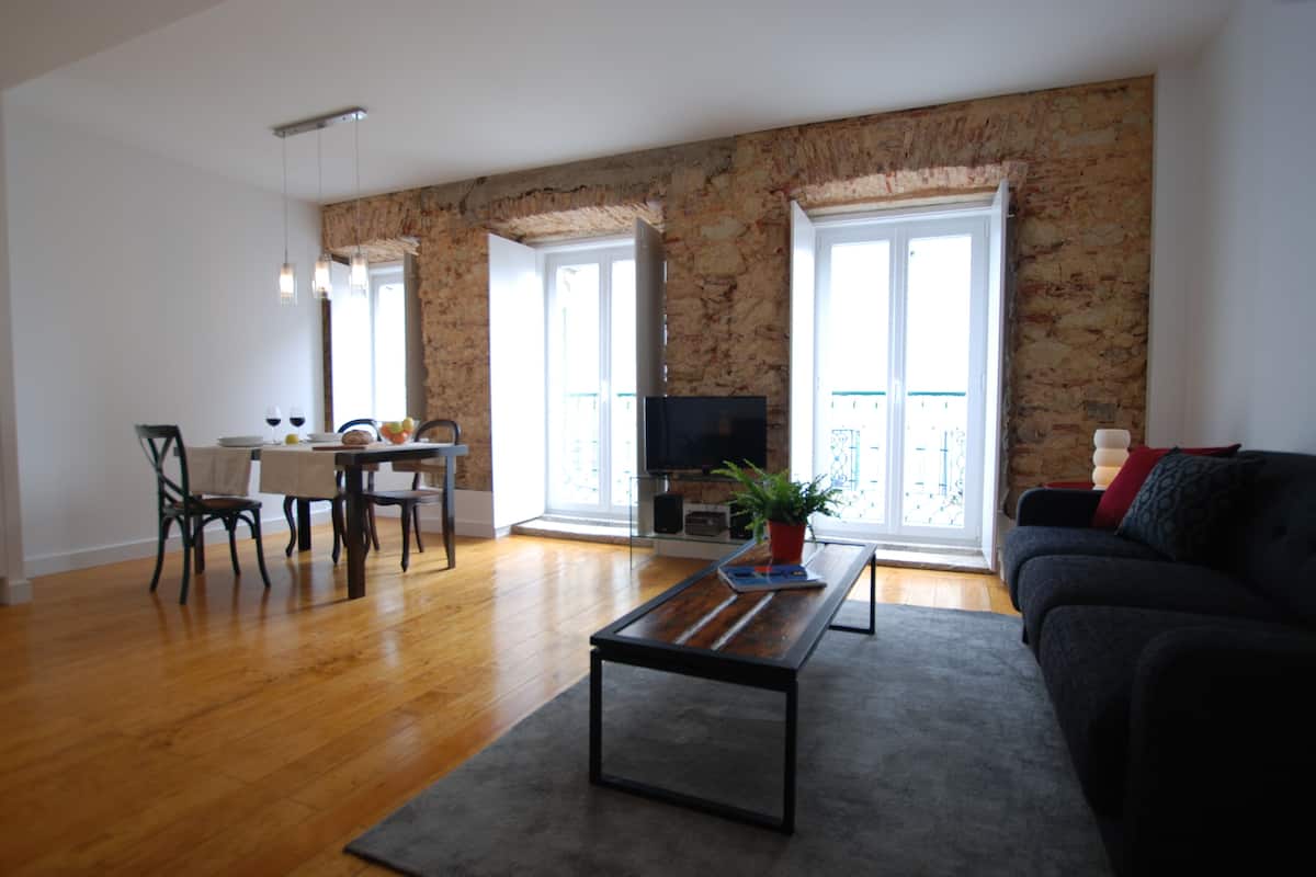 A spacious living area with a wooden floor, a black sofa, a dining table with chairs, and a TV.