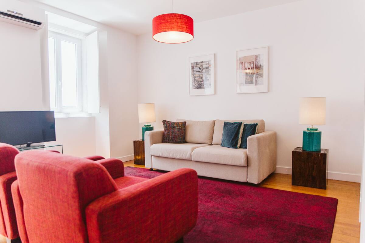 A living room with a sofa, red armchairs, a red rug, a television, and two wall-mounted lamps.