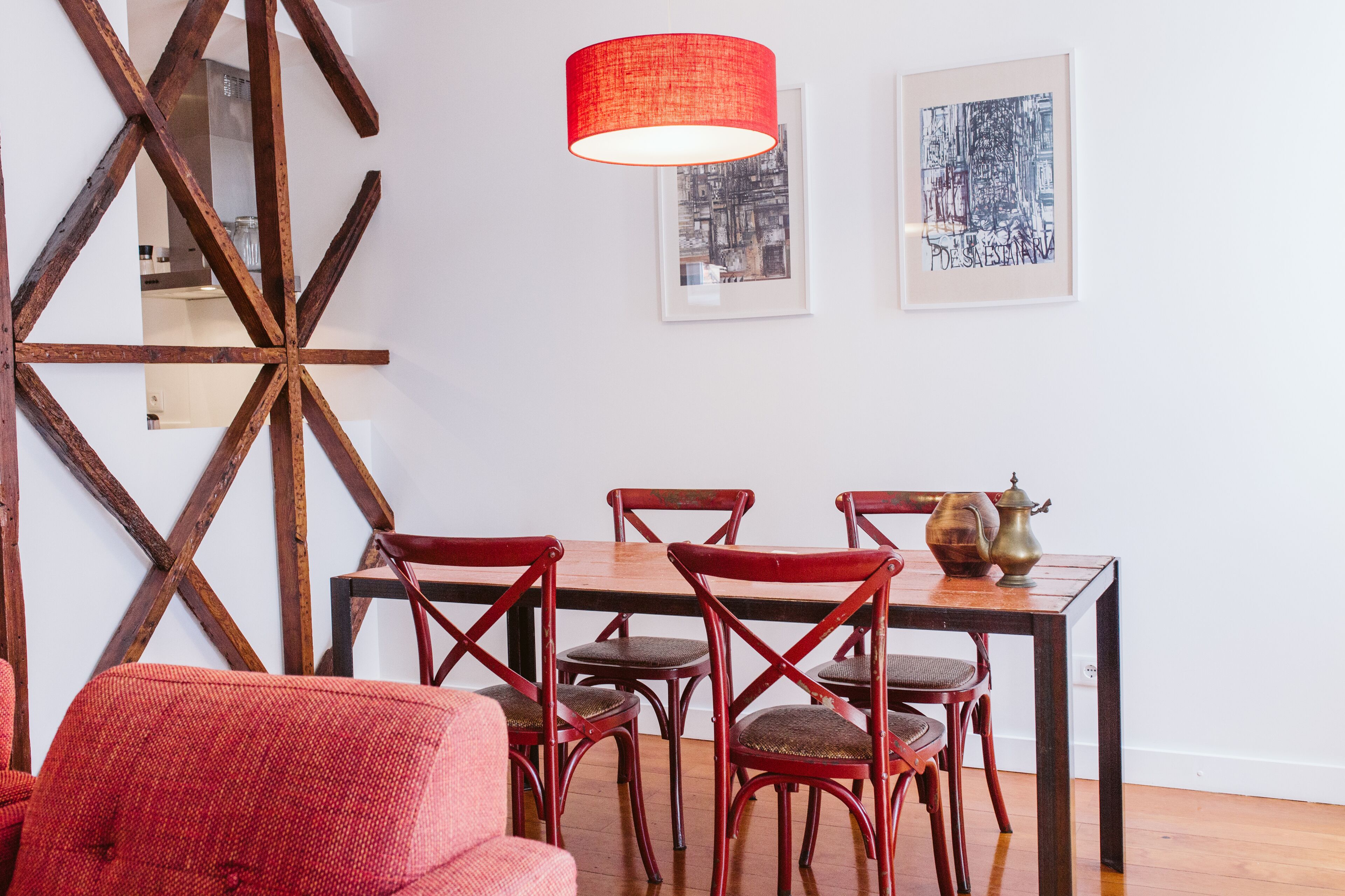 A dining area with a wooden table, red chairs, and a red pendant light. There are framed artworks on the wall.