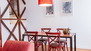 A dining area with a wooden table, red chairs, and a red pendant light. There are framed artworks on the wall.