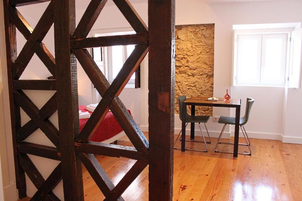 Een kamer met een houten vloer, een stenen muur, een bed met een rode deken, een tafel met stoelen, en een raam.