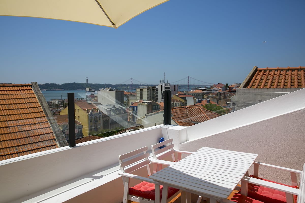 A balcony with a white table and chairs, overlooking a cityscape with buildings and a bridge.