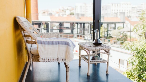 A wicker chair with a white and blue striped blanket, a small wicker table with a vase and wine glasses, and a view of buildings through a window. - 54 Santa Catarina Boutique Aparthotel (Lisbon)