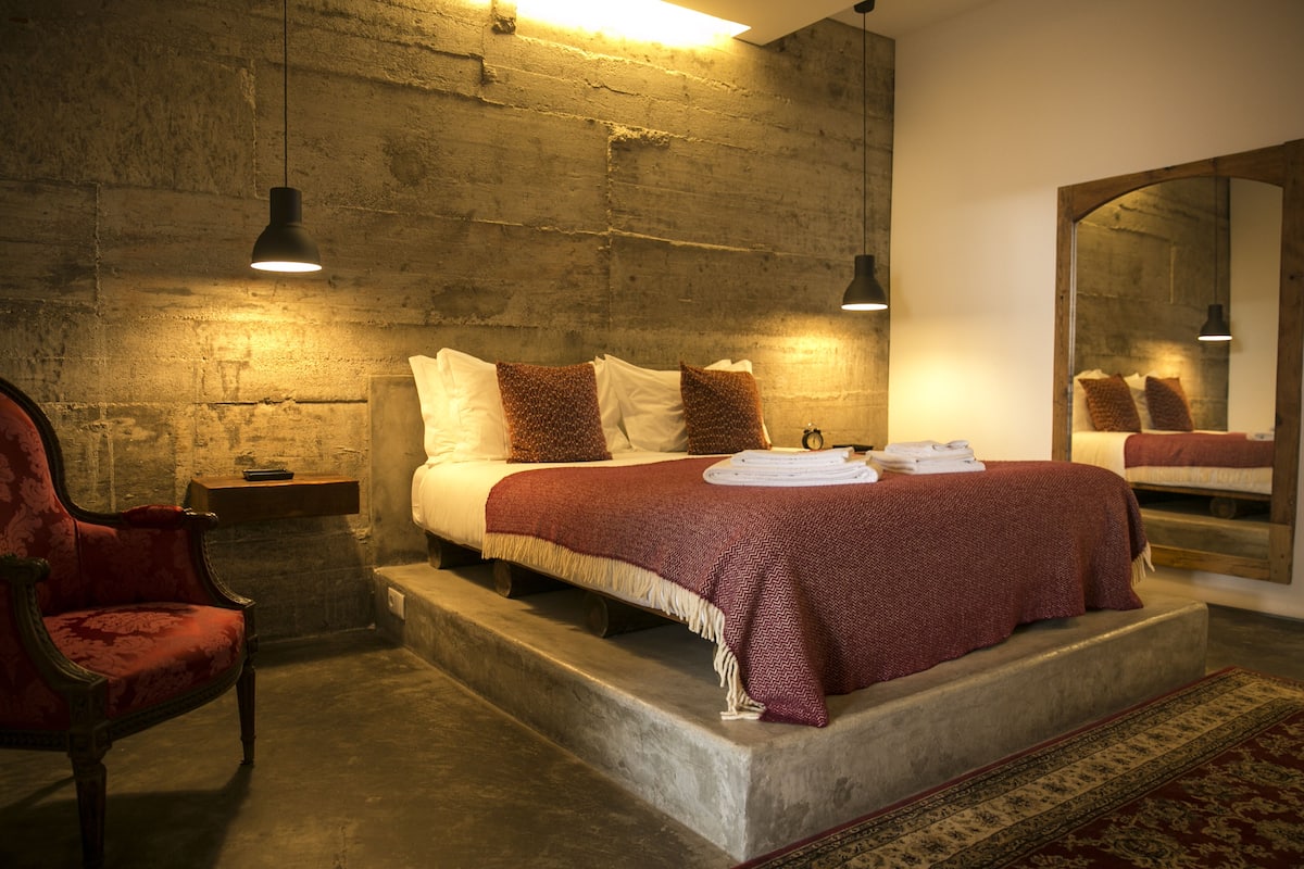 A bedroom with a large bed, a wooden headboard, a red armchair, and a mirror on the wall.