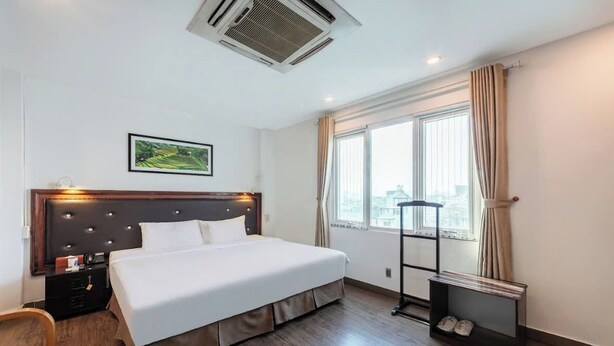 Quarto de hotel com uma cama grande, cabeceira iluminada, janela com cortinas, sapateira e um quadro na parede.