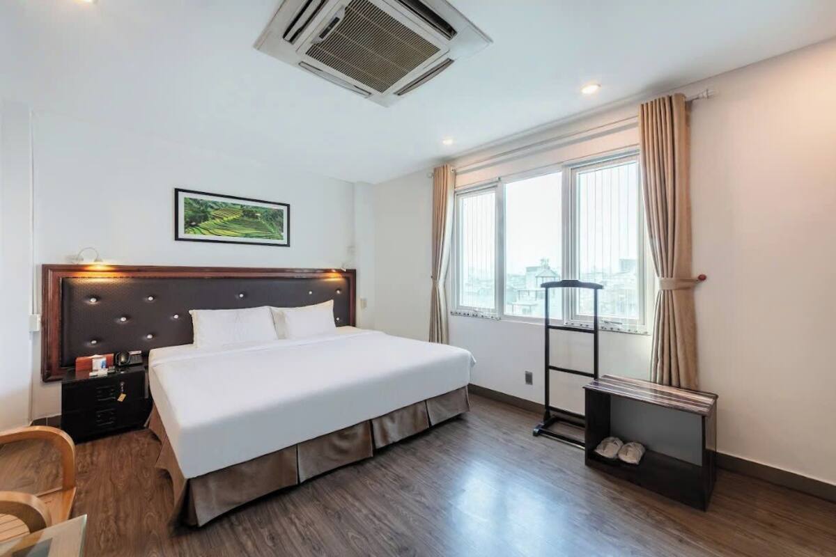 Une chambre d’hôtel avec un grand lit, une tête de lit dotée de lampes, une fenêtre avec des rideaux, un porte-chaussures et un tableau au mur.
