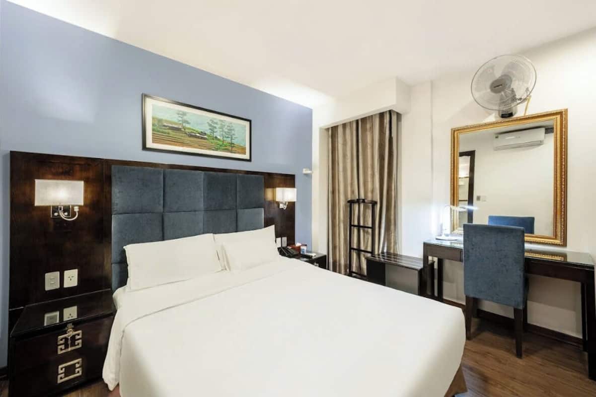 Une chambre d’hôtel avec un grand lit, un bureau avec une chaise, un miroir, un ventilateur et un tableau au mur.