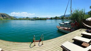 Une terrasse en bois avec des chaises longues et un bateau amarré sur un lac paisible, avec d’autres bateaux en arrière-plan et des montagnes à l’horizon.