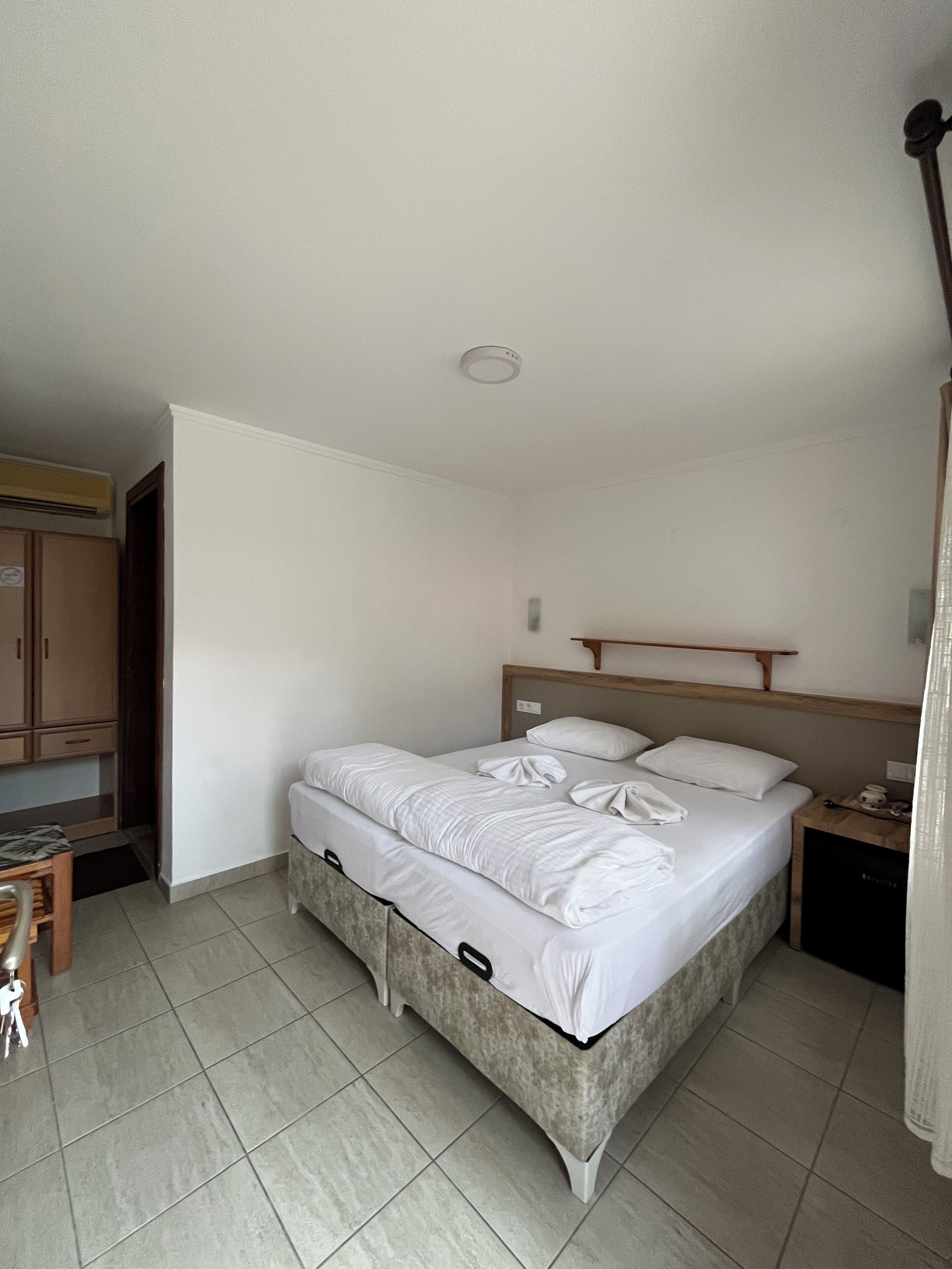 Un lit simple avec du linge de lit blanc, une tête de lit en bois, une table de chevet avec une lampe et une petite armoire.