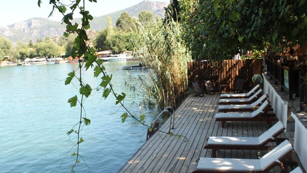 Une terrasse en bois avec des chaises longues, située au bord d’un lac, entourée de verdure et avec des montagnes en arrière-plan.