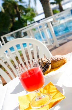 A glass of red drink on a white table with a yellow napkin, next to pastries, in a sunny outdoor setting with a pool and white chairs in the background. - Hotel Metropol (Senigallia)