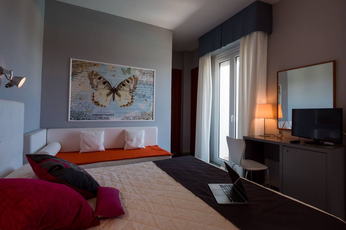 A modern hotel room with a bed, a desk with a laptop, a TV, and a large butterfly artwork on the wall.