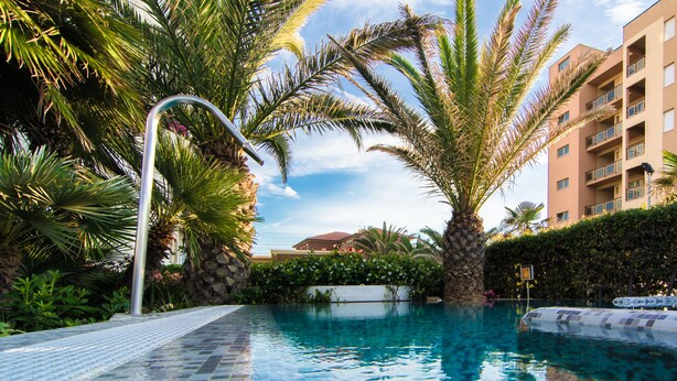 A swimming pool with a tiled deck, surrounded by palm trees and a building in the background. - Hotel Metropol (Senigallia)
